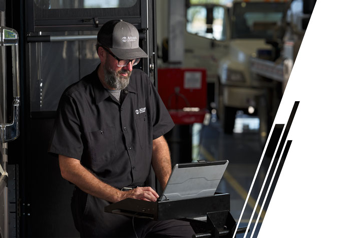 A white man with a gray beard, glasses and a gray hat with "Allison Transmission" on the front works on a computer next to a vehicle.