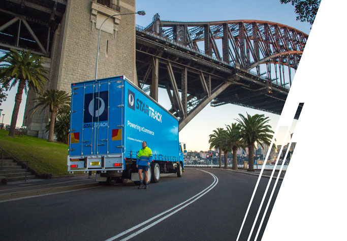 A white box truck with a red cab is parked on the street. A man pushes cardboard boxes on a trolley behind the truck.