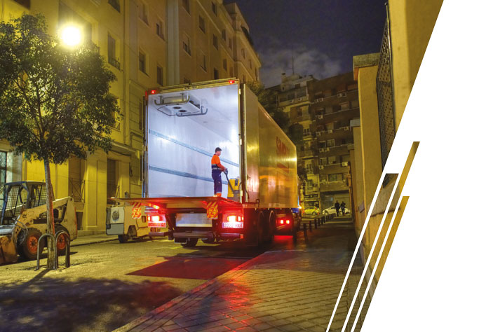 A white box truck with a red cab is parked on the street. A man pushes cardboard boxes on a trolley behind the truck.