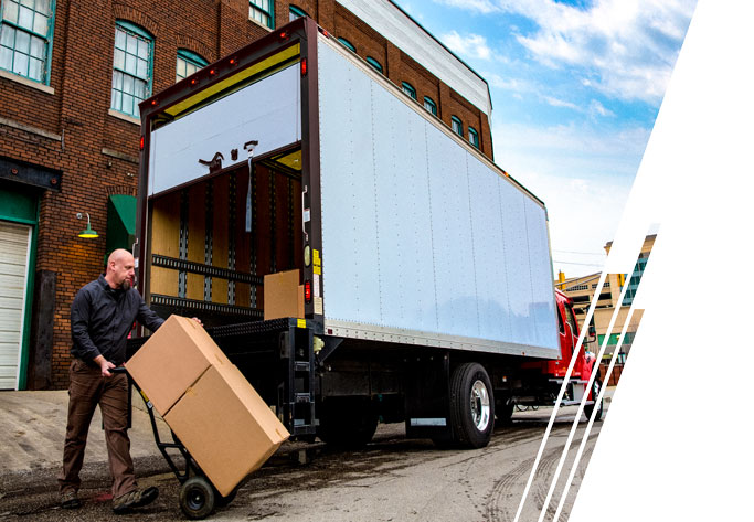 A white box truck with a red cab is parked on the street. A man pushes cardboard boxes on a trolley behind the truck.