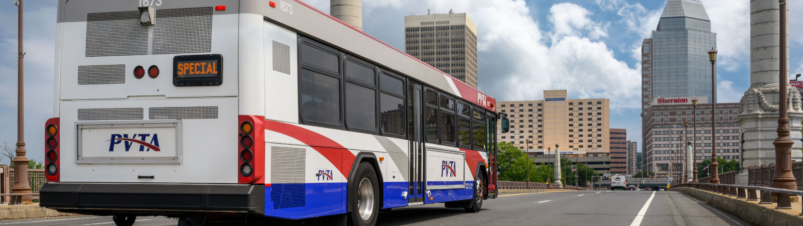 A red, white and blue bus goes across a bridge towards a city.
